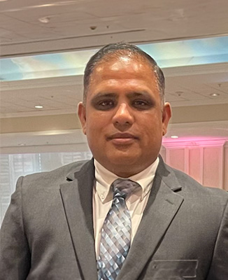 The image shows a man wearing a suit and tie, standing against a backdrop that appears to be an indoor event setting.