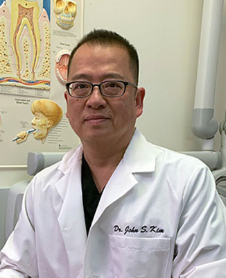 The image is a photograph of a person, likely a medical professional, wearing glasses and a white lab coat with a name tag. They are standing in what appears to be a medical or dental office setting, posing for the camera with a neutral expression.