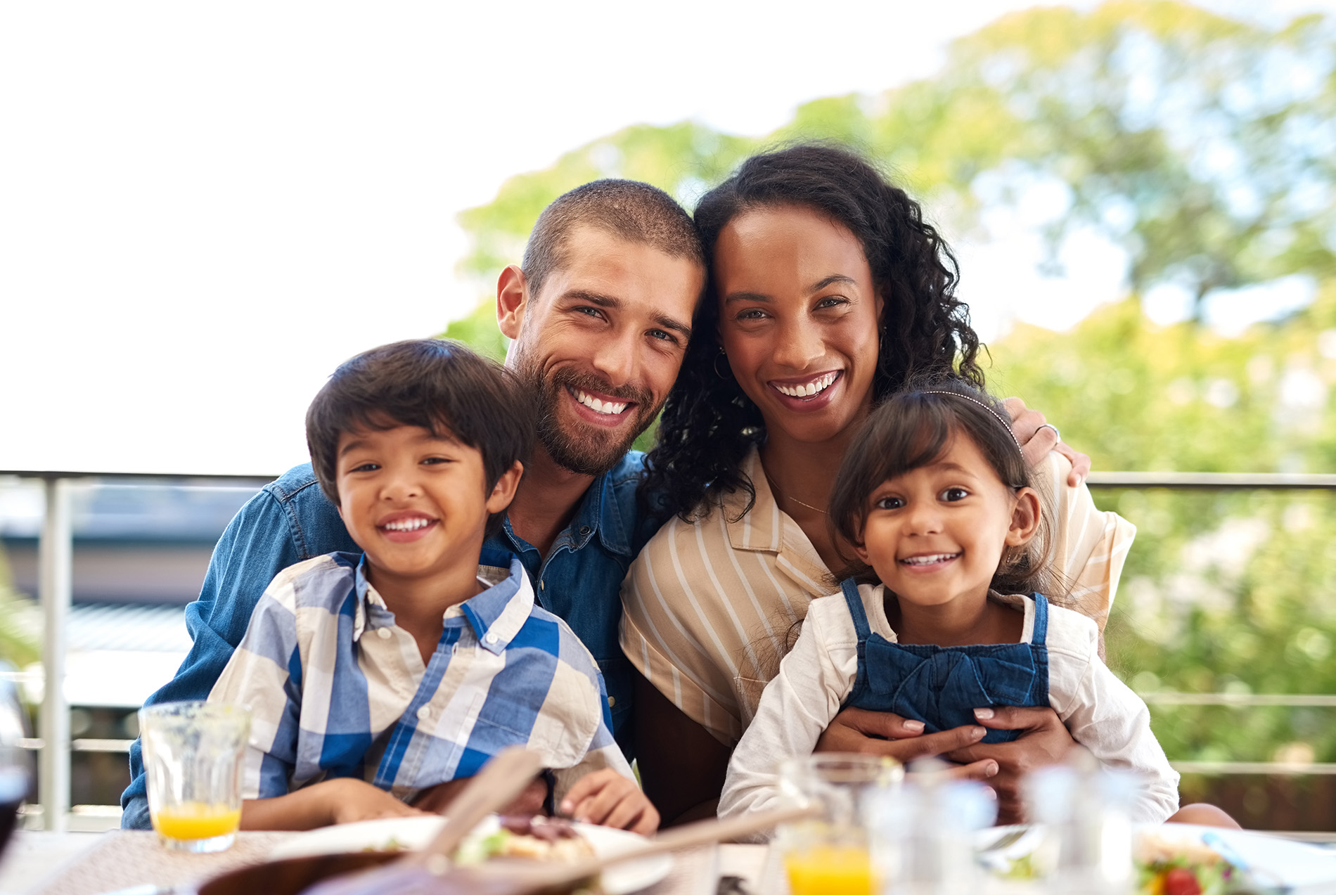 The image shows a family of four, including two adults and two children, posing for a photo at an outdoor dining table during the day.