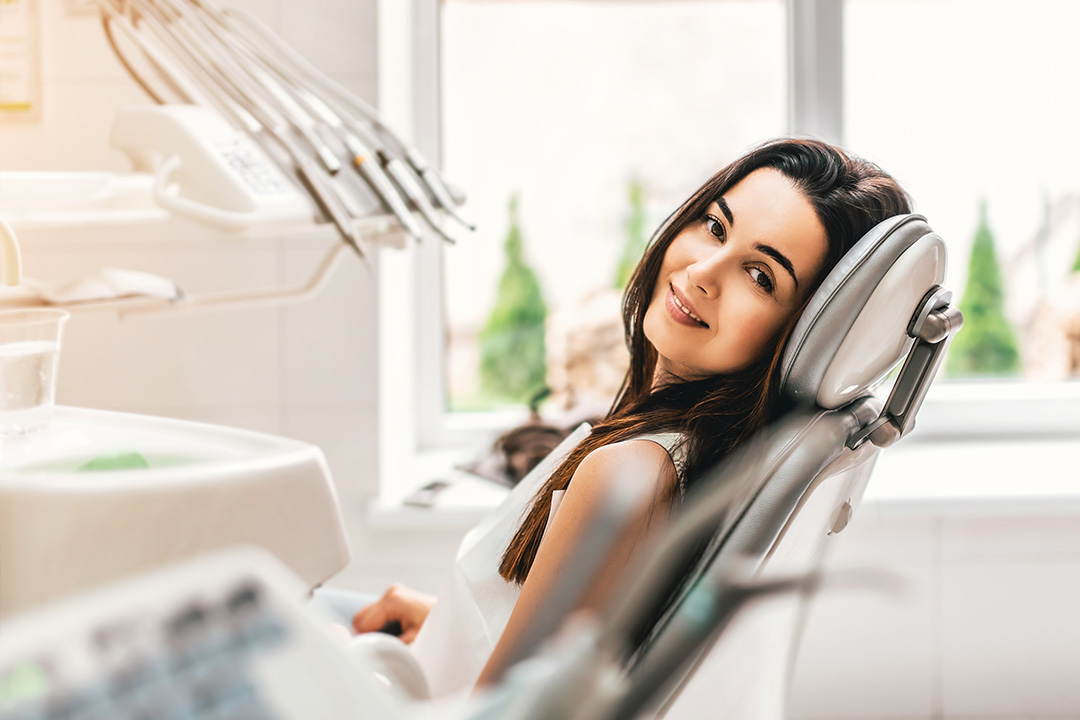 A woman is sitting in a dental chair, smiling at the camera, with dental equipment and a window behind her.