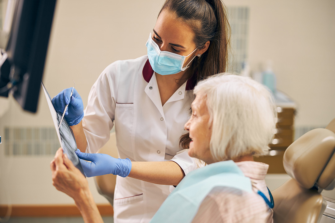 A dentist in a white coat and mask is assisting an elderly patient who is seated in a dental chair, holding a smartphone with both hands.