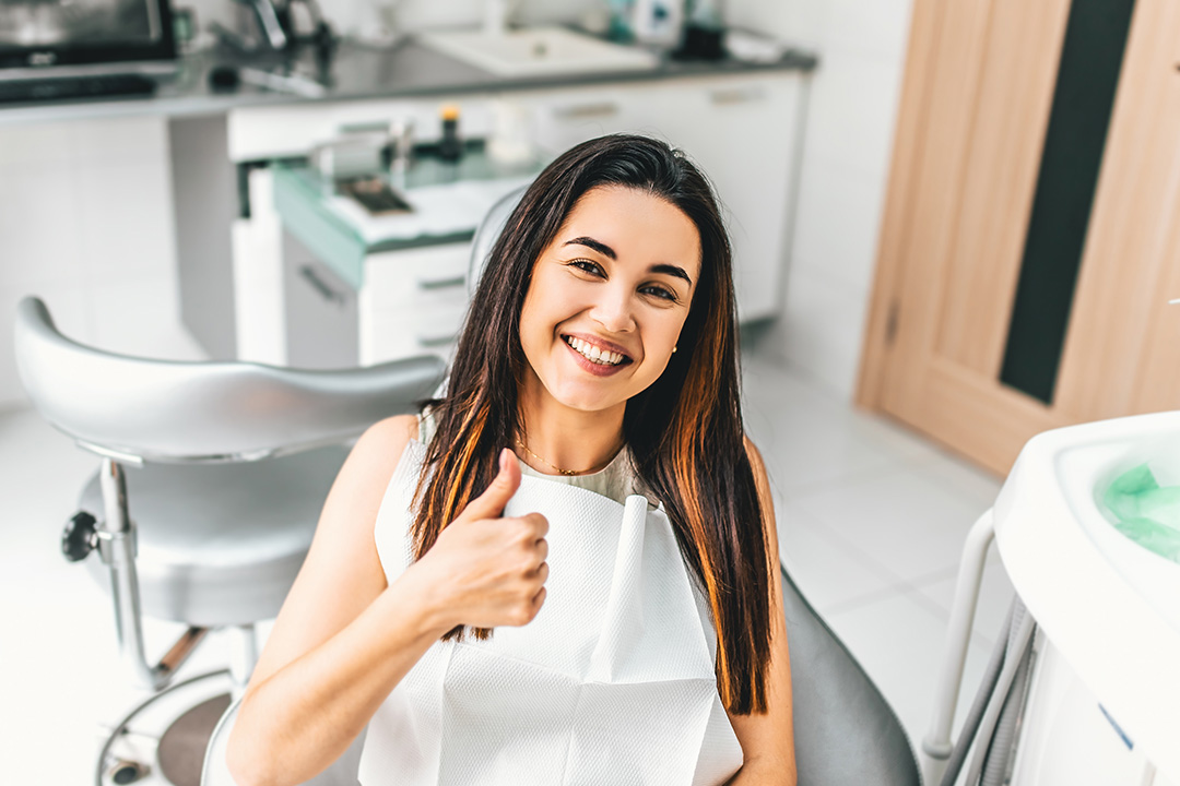 A woman is sitting in a dental chair, smiling and giving a thumbs-up gesture.