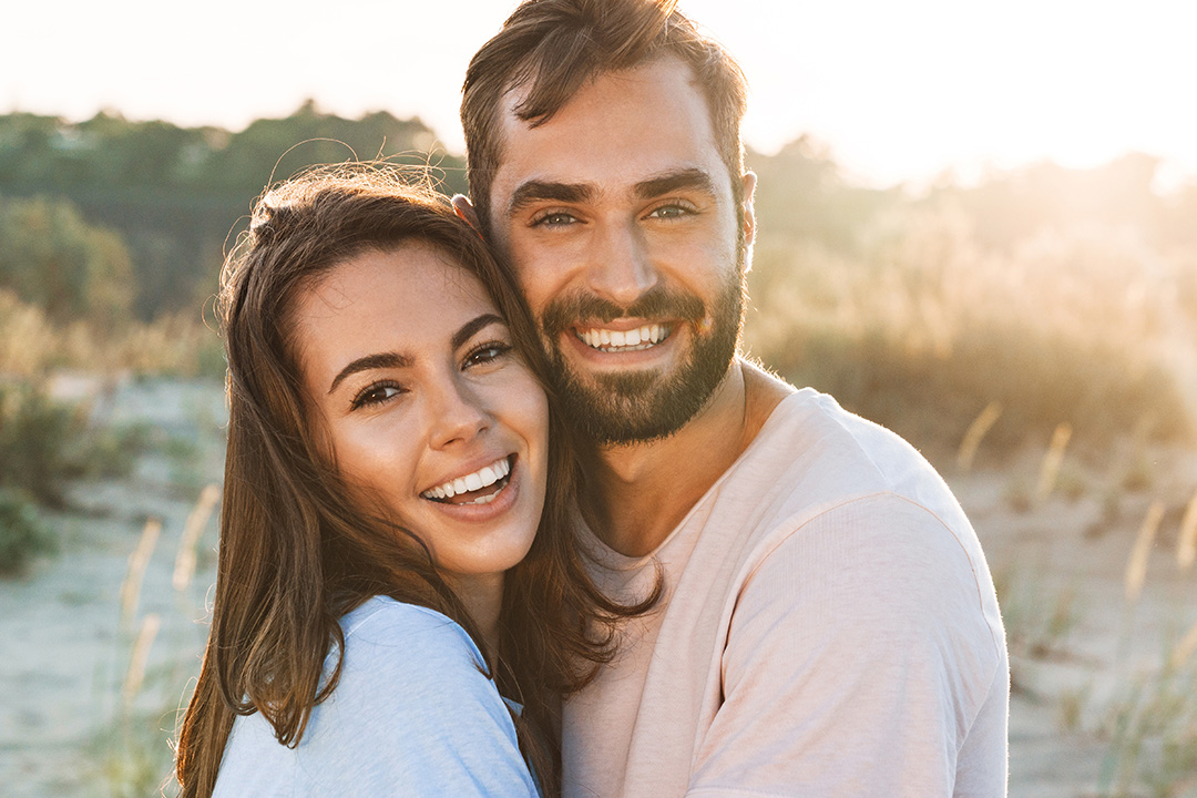 A man and woman smiling at the camera, embracing each other on a beach during sunset.