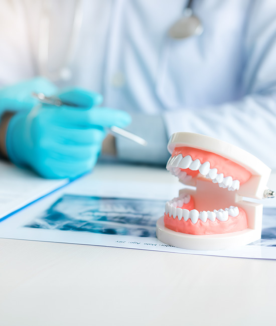 The image shows a healthcare professional, possibly a dentist or oral surgeon, wearing blue gloves and a face mask, sitting at a desk with a patient s dental model in front of them.