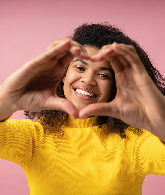 A person is holding their hands together in a heart shape, smiling, and appears to be happy. They are wearing a yellow top and have short hair. The background is a solid color.