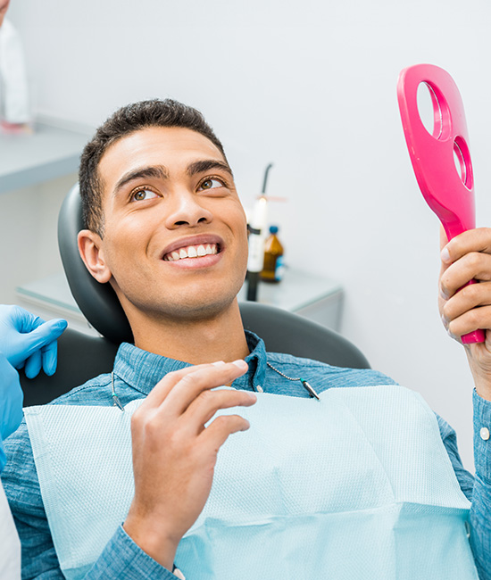 The image shows a dental professional seated in a chair, wearing a blue surgical mask and holding a pink instrument, smiling at the camera.