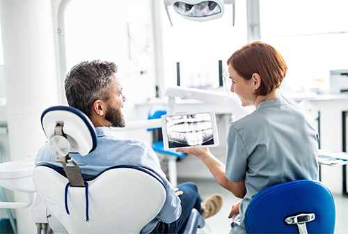 A dental professional is showing a patient an image of their teeth on a tablet while seated in a dental chair, with both wearing face masks.
