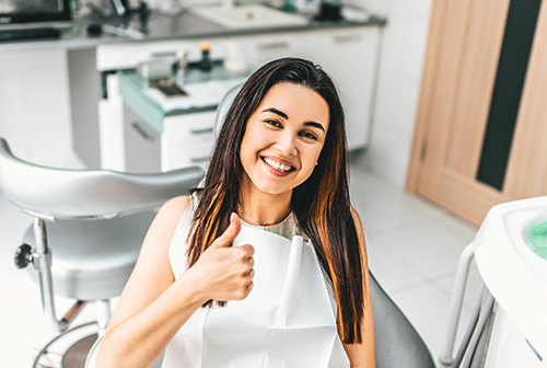 A young woman is sitting in a dental chair, smiling and giving a thumbs-up gesture. She appears to be in a dental office setting.