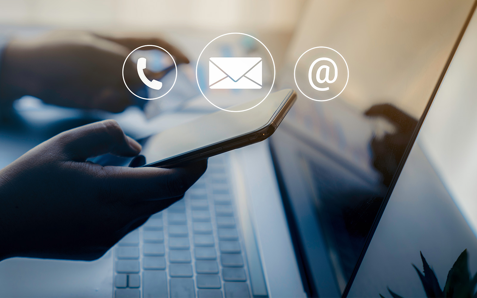 The image shows a person s hand holding a smartphone, displaying an email icon, over a laptop screen with the same email icon.
