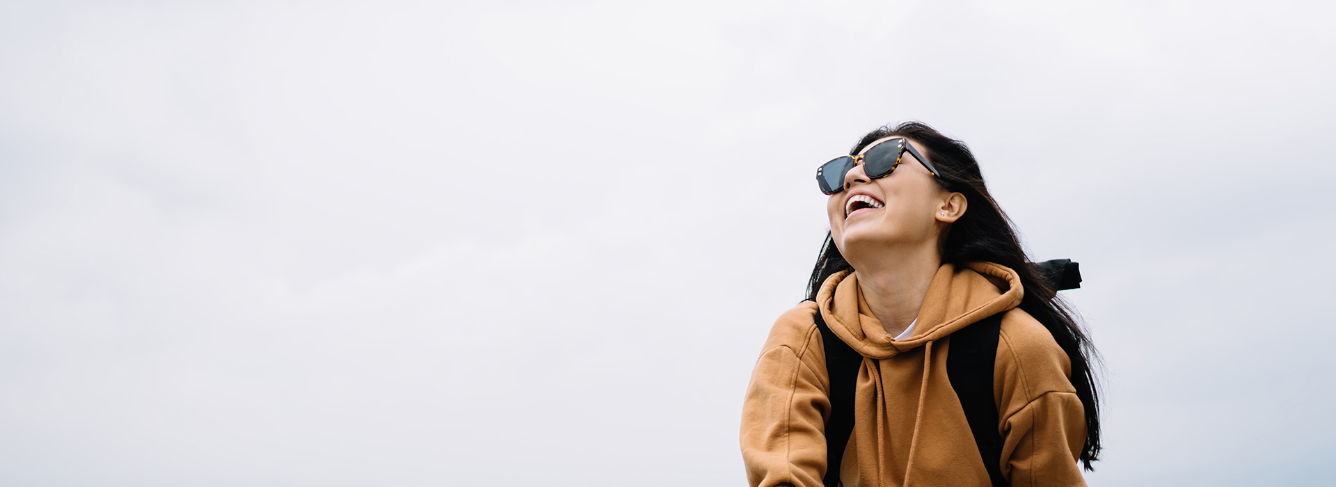 A person is standing outdoors, smiling and looking off to the side while wearing sunglasses and a backpack.