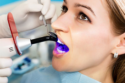 The image shows a woman sitting in a dental chair with a device attached to her mouth, likely for a dental procedure or examination. She is wearing protective eyewear and has a medical mask on her face. A dental professional is performing the procedure, using a device that appears to be a dental scanner or similar diagnostic tool. The environment suggests a professional dental office setting.