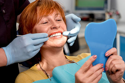 The image shows a smiling woman sitting in a dental chair, holding a blue dental impression tray with both hands, while a dental professional works on her teeth.