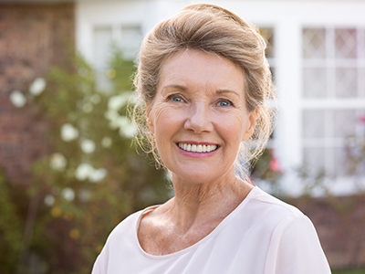 The image is a portrait of an older woman with blonde hair, wearing a white top and smiling at the camera.