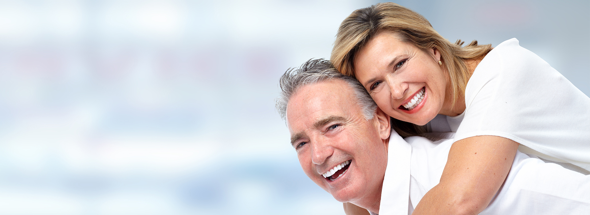 The image is a photograph of a man and woman, presumably in a close relationship, smiling and embracing each other. They appear to be middle-aged adults, with the woman on the left and the man on the right. Both are dressed in light-colored clothing, suggesting a casual or formal setting. The background is blurred but seems to have a gradient of colors that might indicate an indoor environment.