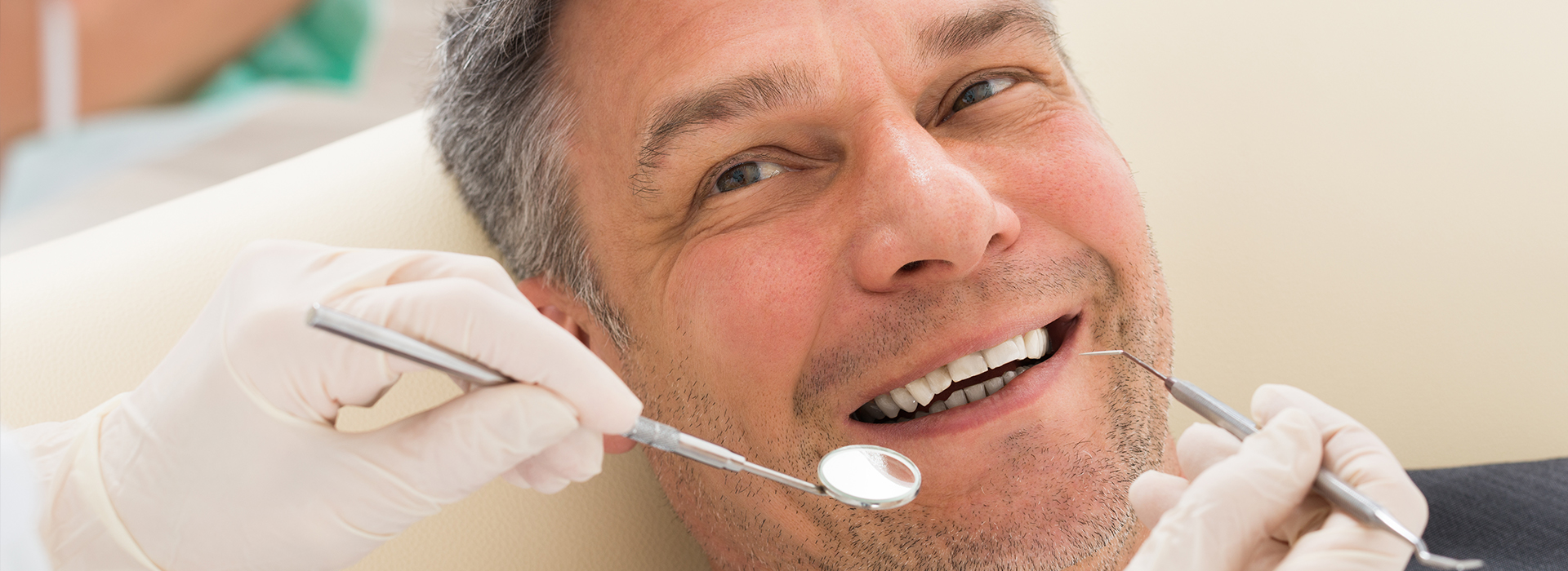 A man sitting in a dental chair, smiling at the camera, with dental instruments and a dentist s hand visible.