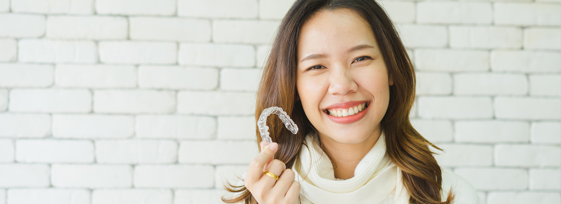 The image shows a smiling woman holding a ring, with a blurred background featuring a brick wall.