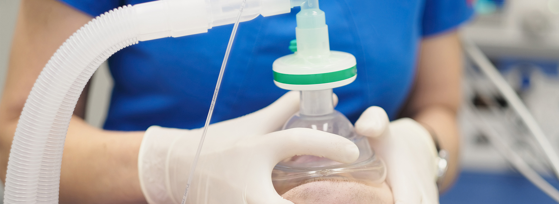 The image shows a person wearing gloves and a blue shirt, holding a clear plastic tube with a green cap connected to a device that appears to be a medical or scientific apparatus.