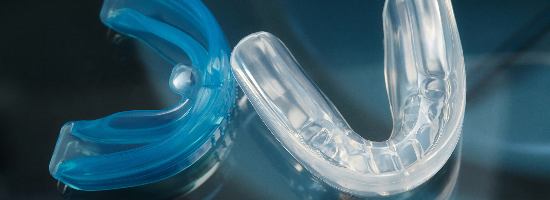 The image shows a close-up of a blue, translucent dental retainer with a clear plastic hook, placed against a blurred background that appears to be a reflective surface.