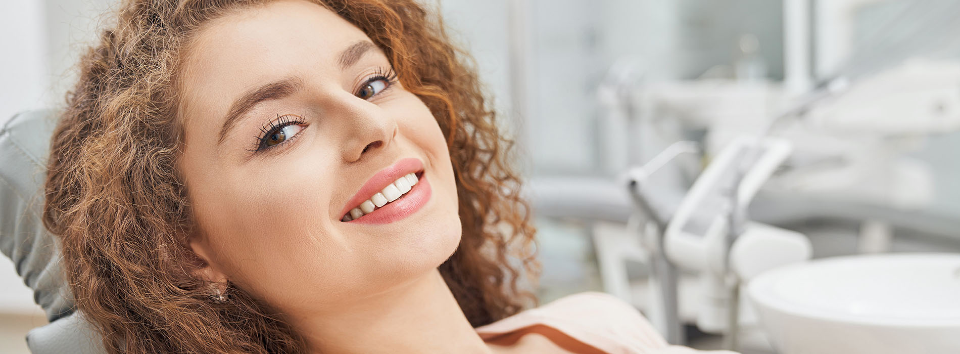 A woman with curly hair, wearing a necklace, smiling at the camera while seated in a dental chair.