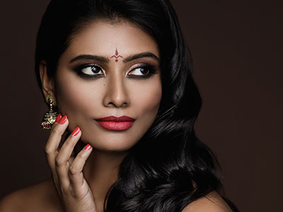 The image features a woman with dark hair and makeup, wearing red nail polish. She is looking directly at the camera with her hand placed on her chin.