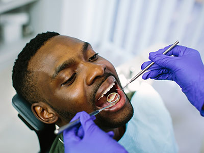 The image shows a person sitting in a dental chair, receiving dental treatment with their mouth open while wearing protective gloves.